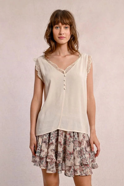 A woman embodies feminine elegance in Molly Bracken's Lace Camisole, a cream sleeveless top with delicate lace trim around the v-neckline and a row of small buttons down the front.