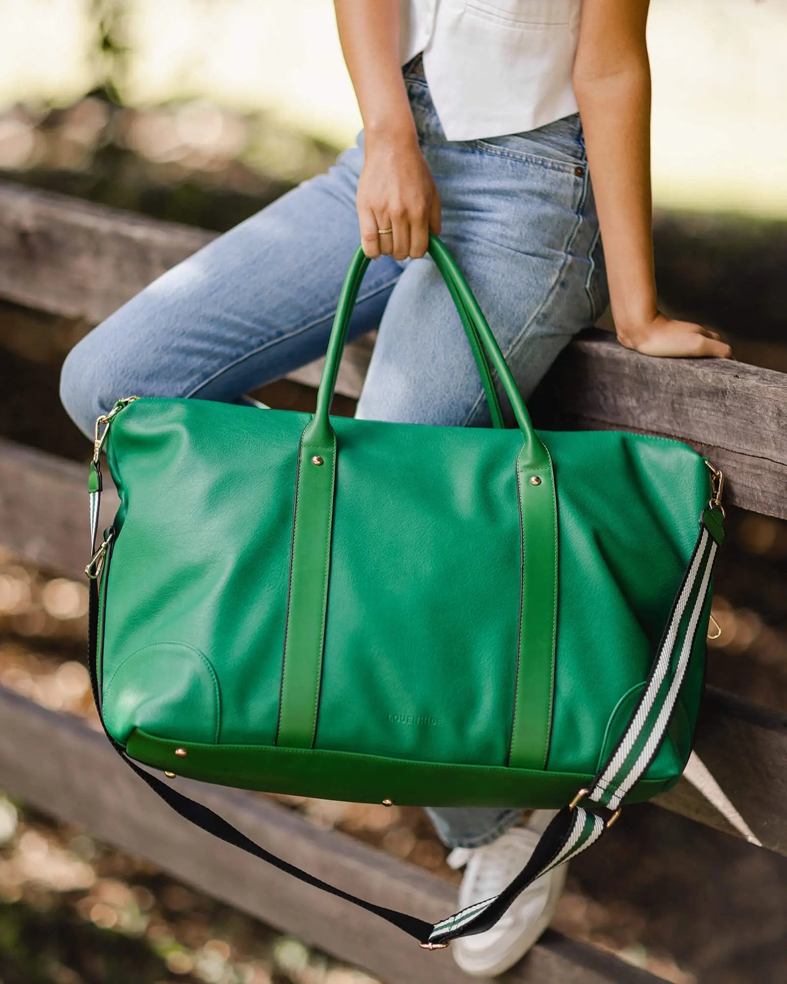 A person holding a large Alexis Stripe Travel Bag by Louenhide, featuring statement stripe webbing and gold hardware, wearing blue jeans.