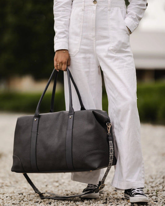 The Alexis Ezra Weekender Travel Bag by Louenhide is a large, dark gray bag made from vegan leather. It features two top handles and an adjustable, patterned shoulder strap.