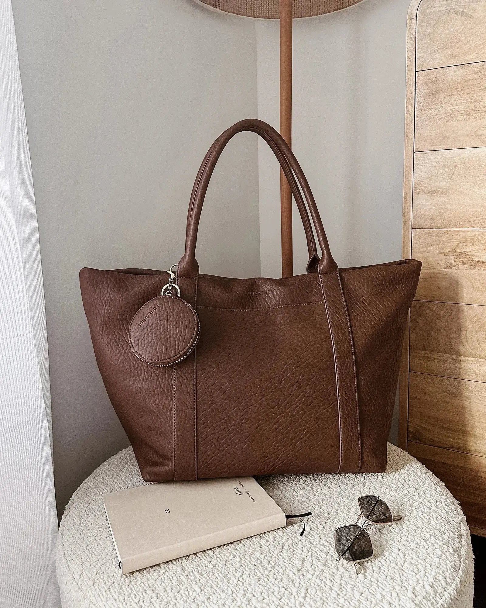 A person in white clothing stands on a porch holding The Savannah Tote Bag by Louenhide, crafted from vegan leather, resting it atop a black rolling suitcase.