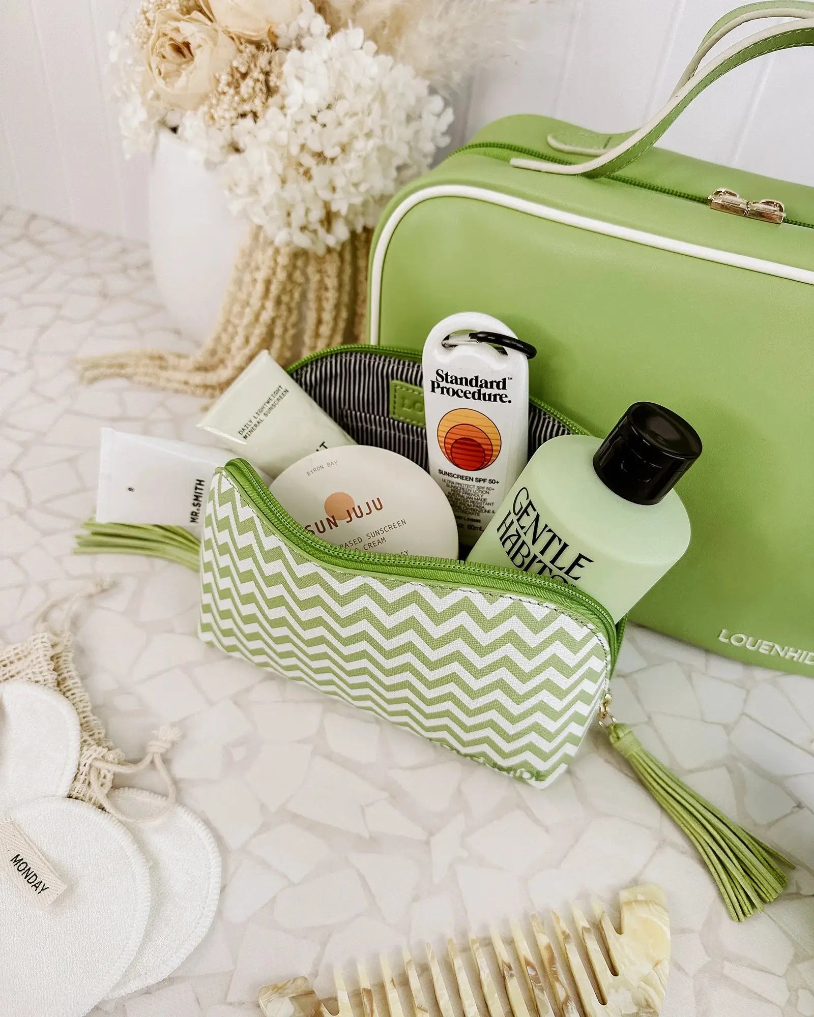 A Louenhide Baby Emma & Audrey Cosmetic Set in green, featuring a front zipper pocket with a partially visible "TO DO" list inside, surrounded by a comb, travel-sized bottle, and white flowers on a tiled surface.