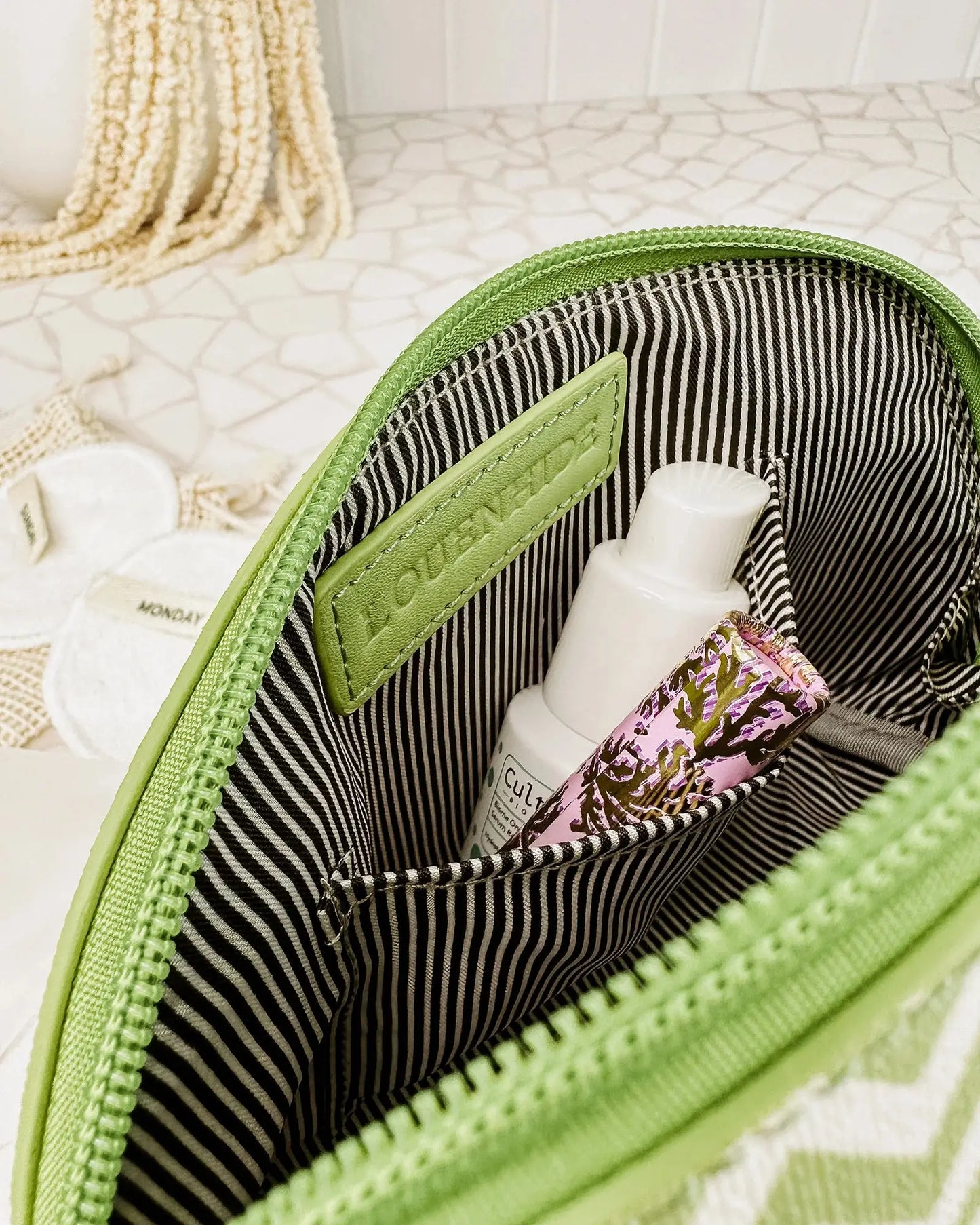 A Louenhide Baby Emma & Audrey Cosmetic Set in green, featuring a front zipper pocket with a partially visible "TO DO" list inside, surrounded by a comb, travel-sized bottle, and white flowers on a tiled surface.