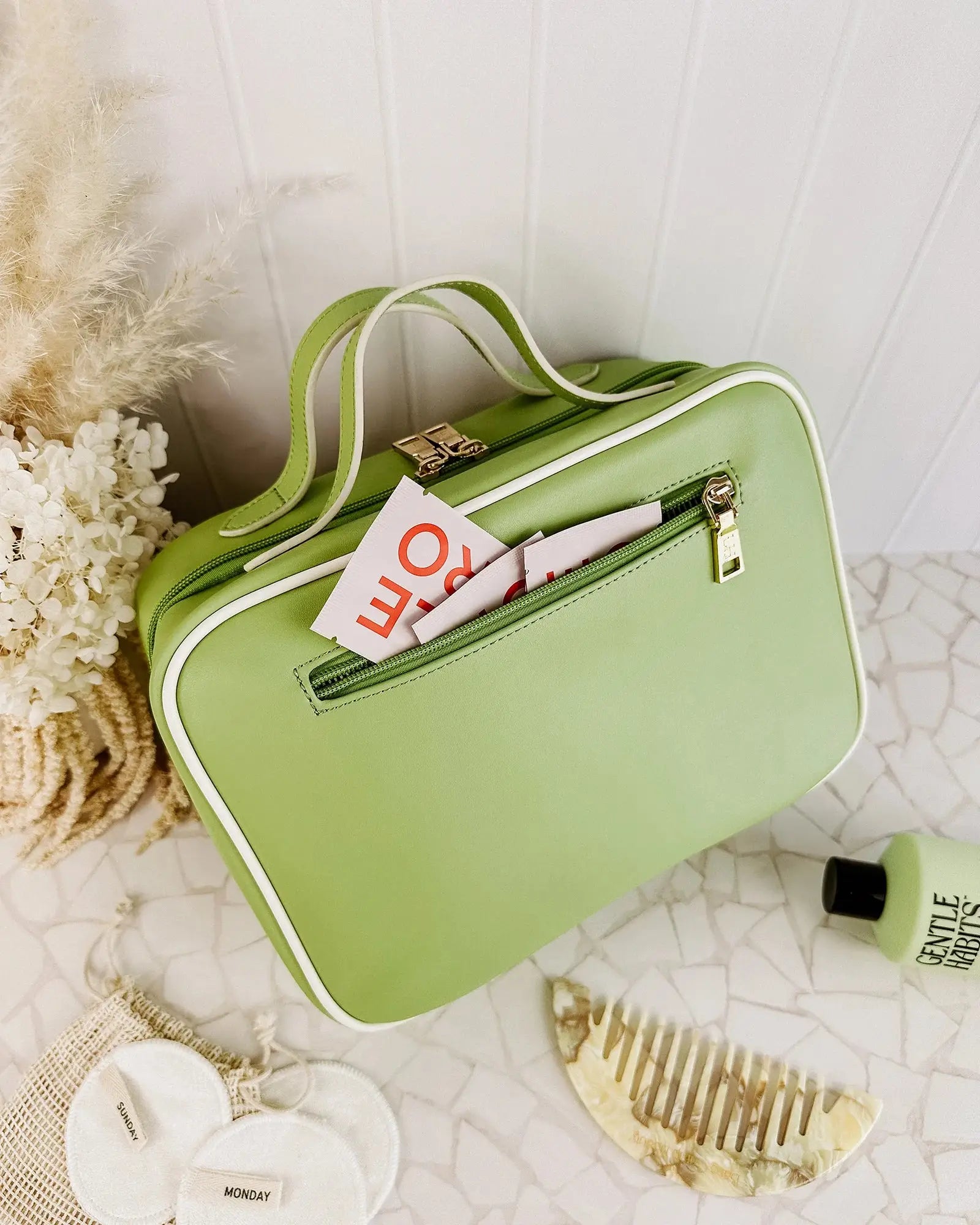 A Louenhide Baby Emma & Audrey Cosmetic Set in green, featuring a front zipper pocket with a partially visible "TO DO" list inside, surrounded by a comb, travel-sized bottle, and white flowers on a tiled surface.