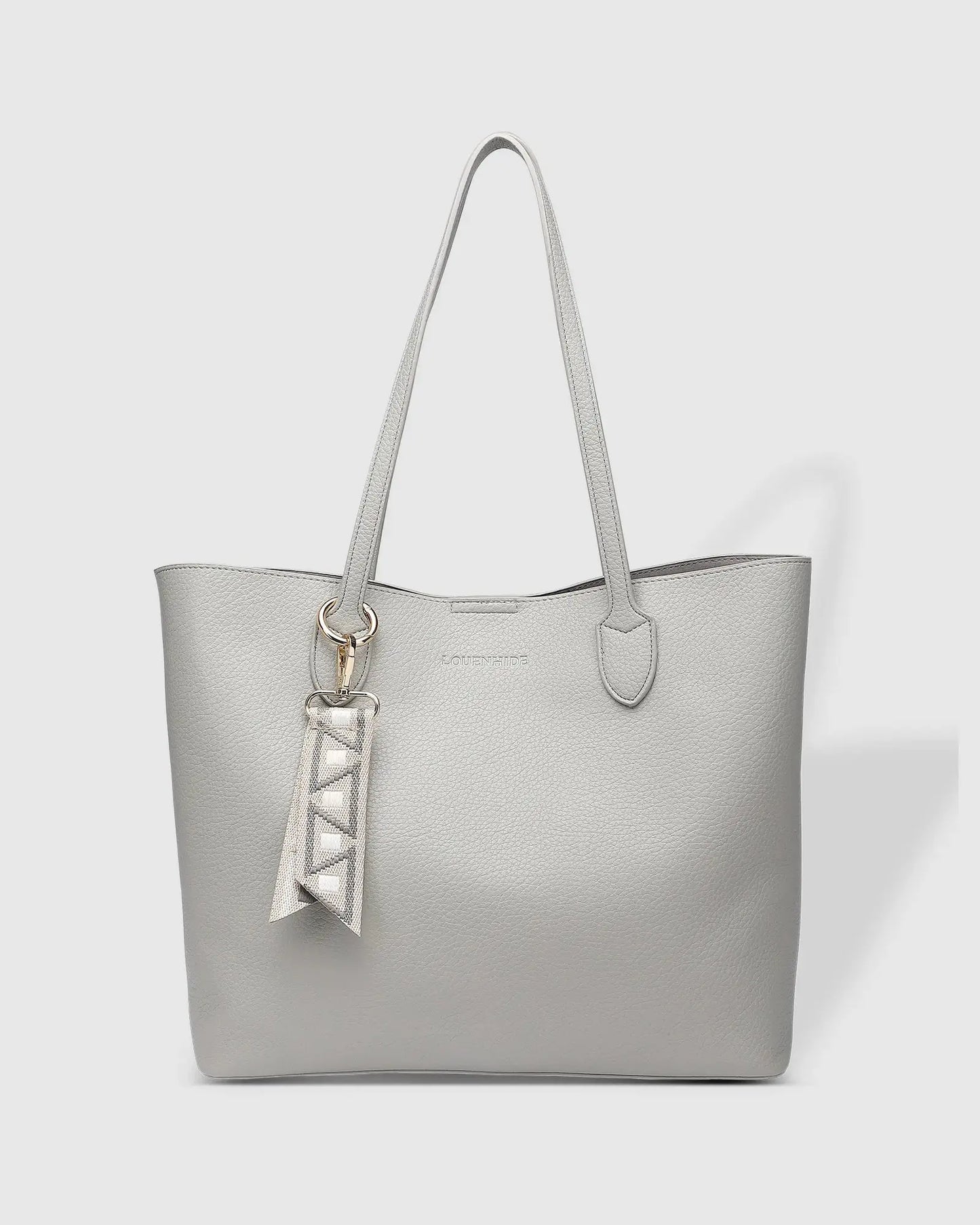 A person wearing a black blazer and white dress, holding Louenhide's The Brea Tote Bag in light grey with a small ribbon detail, stands outdoors near a brick wall with greenery in the background.