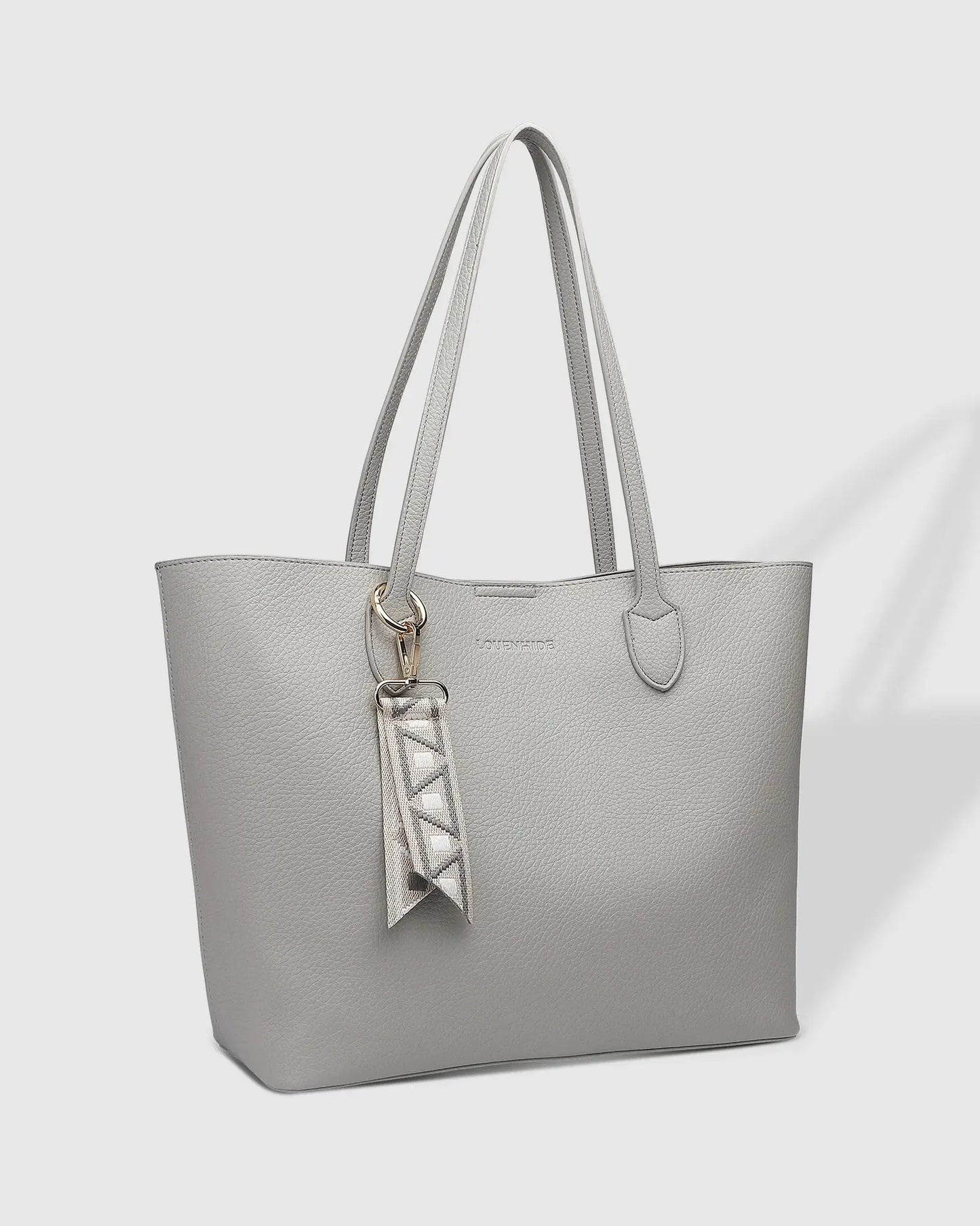 A person wearing a black blazer and white dress, holding Louenhide's The Brea Tote Bag in light grey with a small ribbon detail, stands outdoors near a brick wall with greenery in the background.