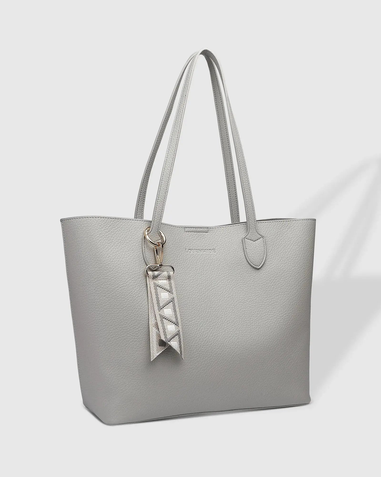 A person wearing a black blazer and white dress, holding Louenhide's The Brea Tote Bag in light grey with a small ribbon detail, stands outdoors near a brick wall with greenery in the background.