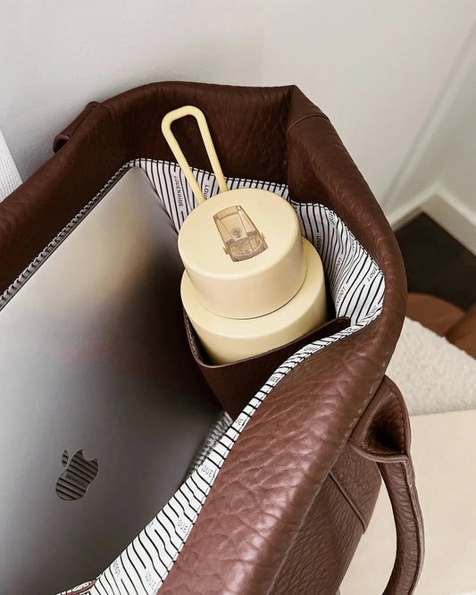 A person in white clothing stands on a porch holding The Savannah Tote Bag by Louenhide, crafted from vegan leather, resting it atop a black rolling suitcase.
