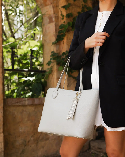 A person wearing a black blazer and white dress, holding Louenhide's The Brea Tote Bag in light grey with a small ribbon detail, stands outdoors near a brick wall with greenery in the background.