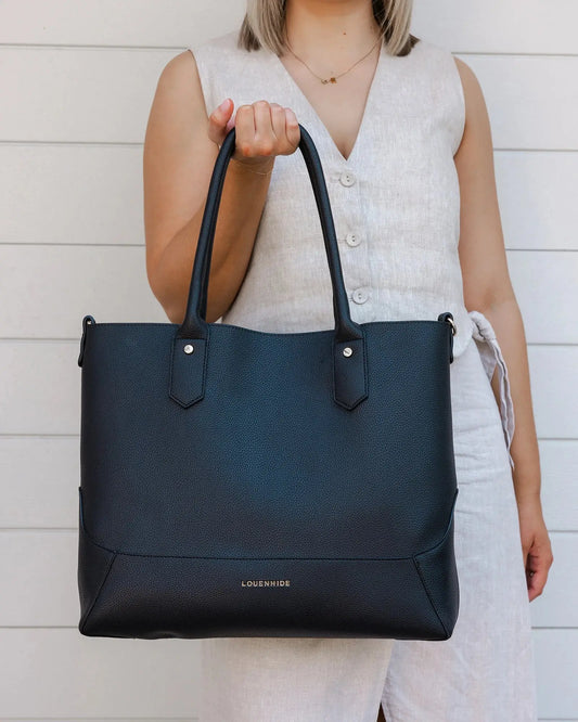 A person with blonde hair in a white outfit poses against a white wall, holding a large black Portsea Tote Bag from Louenhide made of vegan leather over their shoulder.