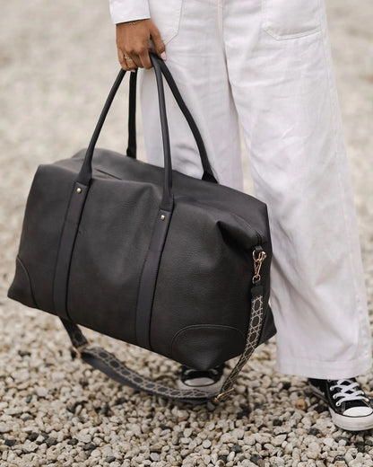 The Alexis Ezra Weekender Travel Bag by Louenhide is a large, dark gray bag made from vegan leather. It features two top handles and an adjustable, patterned shoulder strap.