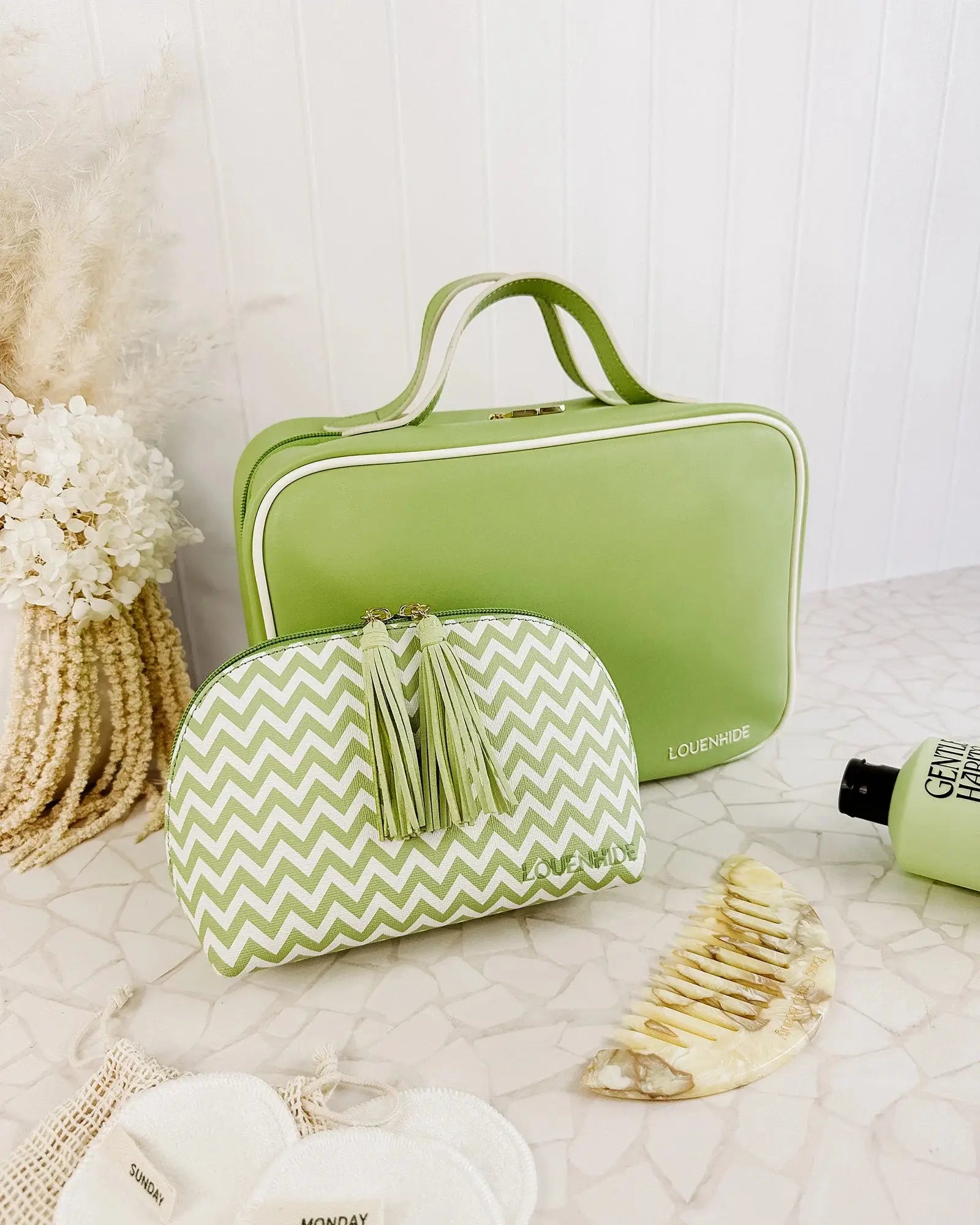 A Louenhide Baby Emma & Audrey Cosmetic Set in green, featuring a front zipper pocket with a partially visible "TO DO" list inside, surrounded by a comb, travel-sized bottle, and white flowers on a tiled surface.