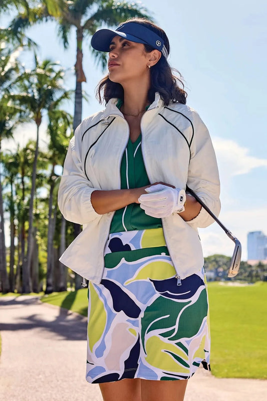 A woman in a white jacket and Tribal HIGH PERFORMANCE PULL-ON SKORT, with a visor and golf glove, stands on a golf course holding a club amid palm trees and buildings.