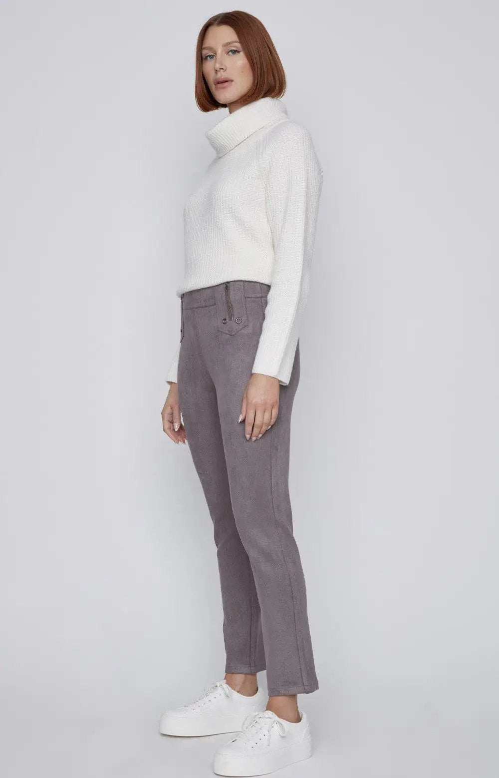 A person with short red hair is standing against a plain background, wearing a white turtleneck sweater, CoCo Y Club gray Front Zip Detail Pant, and white sneakers.