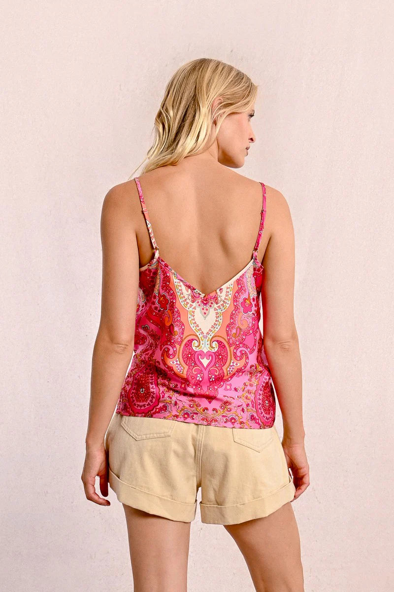 A person with long blonde hair sits on the floor against a plain background, wearing a stylish and comfortable pink patterned top from Molly Bracken called the Backless Camisole, paired with beige shorts.