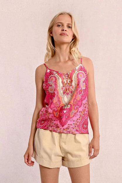 A person with long blonde hair sits on the floor against a plain background, wearing a stylish and comfortable pink patterned top from Molly Bracken called the Backless Camisole, paired with beige shorts.