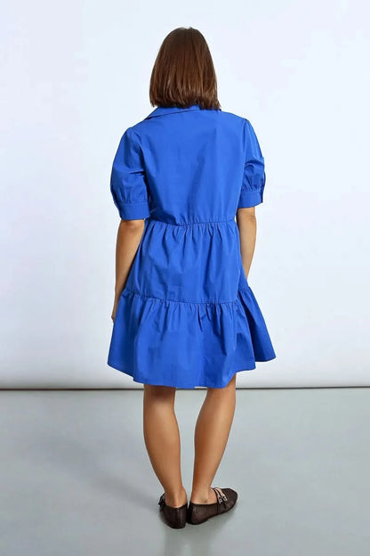 A woman stands elegantly in a Molly Bracken Ruffled Shirt Dress, tiered and blue, paired with black shoes, her hands resting by her sides against a plain background.