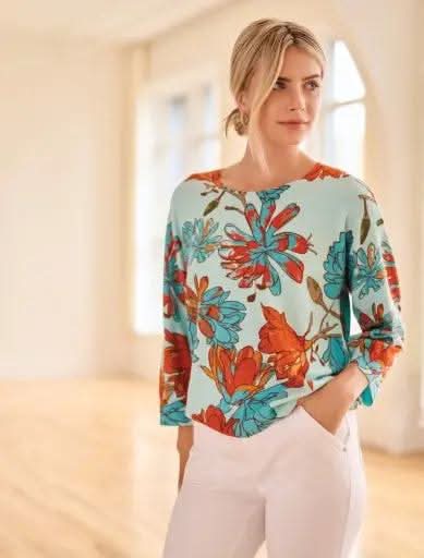 A woman stands indoors in Tribal’s 3/4 Sleeve Crew Neck Sweater—a loose, high-quality top in light blue with red and orange floral print—paired with white pants for a chic, modern look.