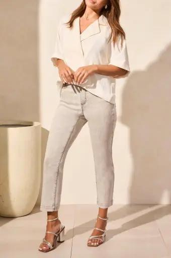 A woman stands indoors wearing the Tribal FLOWY POPOVER SHORT SLEEVE SHIRT with light cropped pants and white heeled sandals. A large cream-colored planter is visible in the background.