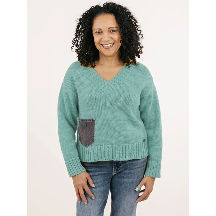 A smiling woman with curly hair wears the Anniston Pullover by Shannon Passero—an ethically made, handloomed teal sweater with a gray pocket—and blue jeans, standing against a plain light background.