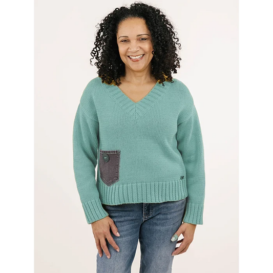 A smiling woman with curly hair wears the Anniston Pullover by Shannon Passero—an ethically made, handloomed teal sweater with a gray pocket—and blue jeans, standing against a plain light background.