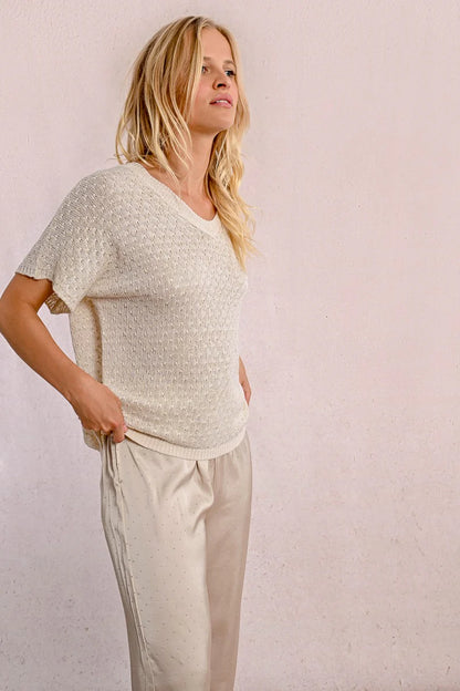 A person with long blond hair stands against a light wall, hands in pockets, wearing the Short Sleeve Knit Top by Molly Bracken and loose pants.