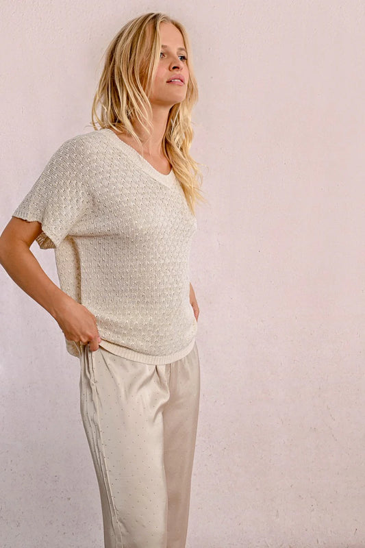 A person with long blond hair stands against a light wall, hands in pockets, wearing the Short Sleeve Knit Top by Molly Bracken and loose pants.