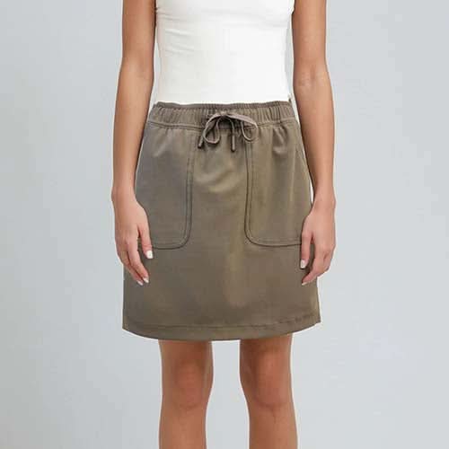 A woman wears the Point Zero Skort—a khaki athletic skort with front pockets and built-in shorts—paired with a white sleeveless top, standing against a plain light background.