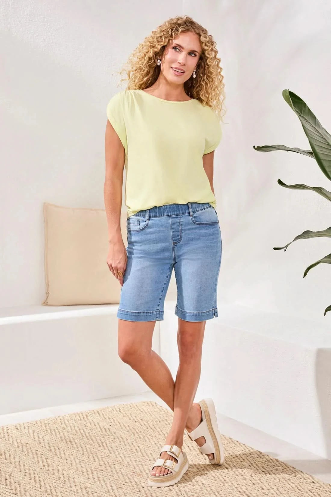 A person stands indoors on a woven rug near a white bench with a beige pillow, wearing Tribal's AUDREY PULL-ON BERMUDA SHORT in light blue stretch denim, a pale yellow top, and beige sandals.