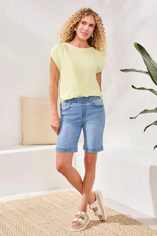 A person stands indoors on a woven rug near a white bench with a beige pillow, wearing Tribal's AUDREY PULL-ON BERMUDA SHORT in light blue stretch denim, a pale yellow top, and beige sandals.