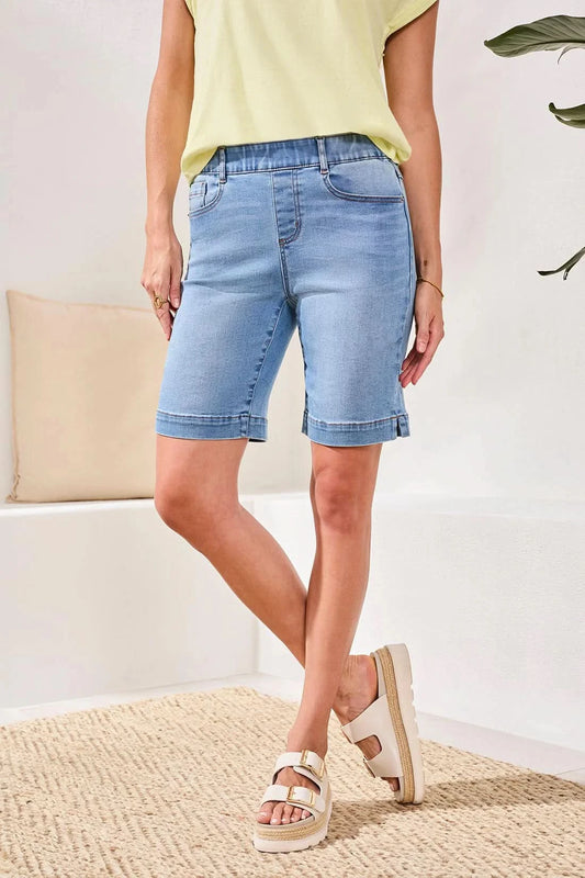 A person stands indoors on a woven rug near a white bench with a beige pillow, wearing Tribal's AUDREY PULL-ON BERMUDA SHORT in light blue stretch denim, a pale yellow top, and beige sandals.