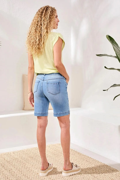 A person stands indoors on a woven rug near a white bench with a beige pillow, wearing Tribal's AUDREY PULL-ON BERMUDA SHORT in light blue stretch denim, a pale yellow top, and beige sandals.