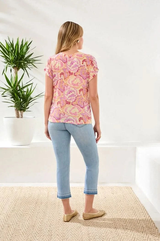 A woman stands indoors on a woven rug, wearing the Tribal CAP SLEEVE WEAR TWO WAYS TOP in a pink pattern with light blue cropped jeans and beige woven shoes. Two potted plants sit in the background.