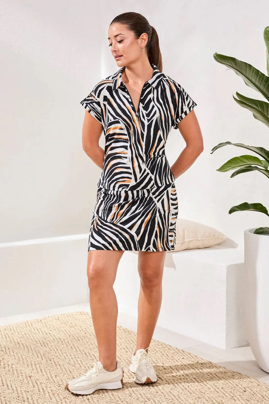 A woman indoors wears the Tribal DOLMAN ZIP-FRONT SPORT TOP with white sneakers and her hair in a ponytail. A large potted plant and white cushion are seen in the background.