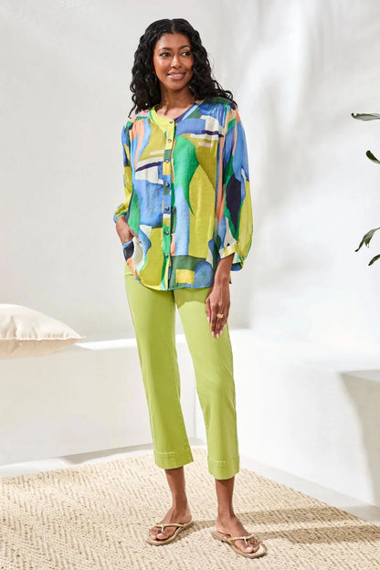 A woman stands indoors wearing the Tribal FLOWY DOLMAN SLEEVE BLOUSE, paired with light green cropped pants and tan sandals, with one hand in her pocket.