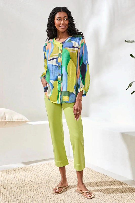 A woman stands indoors wearing the Tribal FLOWY DOLMAN SLEEVE BLOUSE, paired with light green cropped pants and tan sandals, with one hand in her pocket.