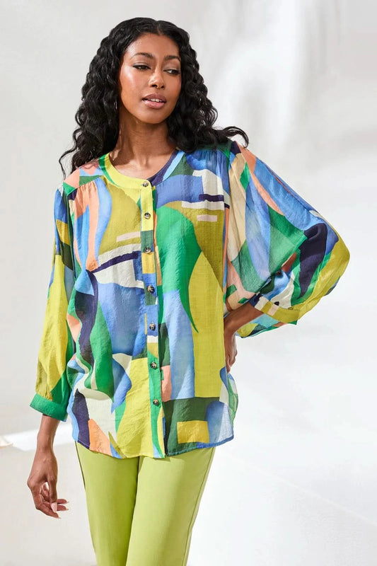 A woman stands indoors wearing the Tribal FLOWY DOLMAN SLEEVE BLOUSE, paired with light green cropped pants and tan sandals, with one hand in her pocket.
