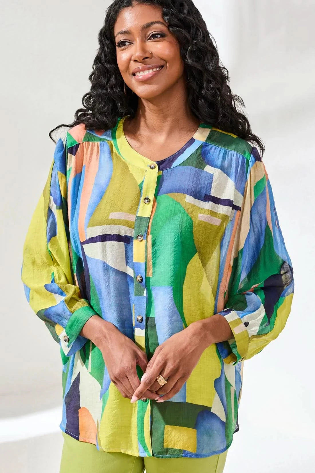 A woman stands indoors wearing the Tribal FLOWY DOLMAN SLEEVE BLOUSE, paired with light green cropped pants and tan sandals, with one hand in her pocket.