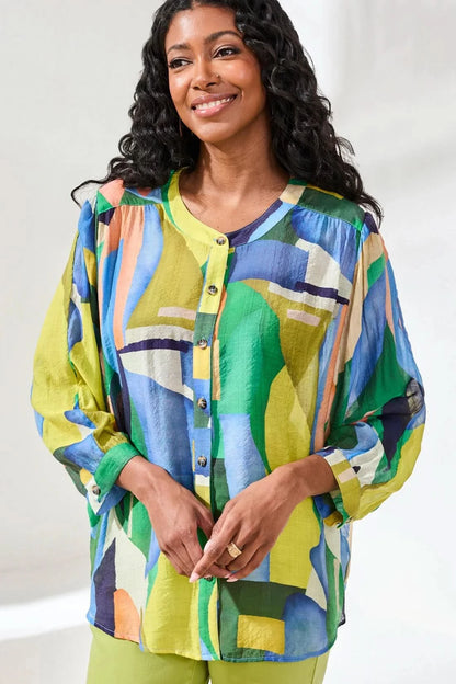 A woman stands indoors wearing the Tribal FLOWY DOLMAN SLEEVE BLOUSE, paired with light green cropped pants and tan sandals, with one hand in her pocket.