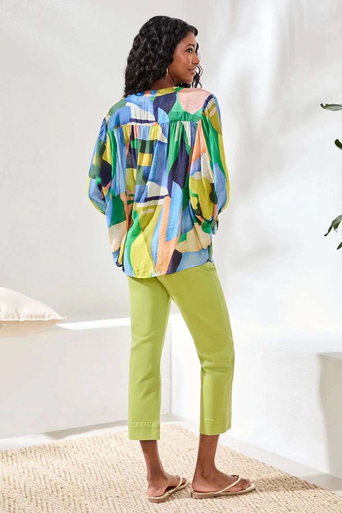 A woman stands indoors wearing the Tribal FLOWY DOLMAN SLEEVE BLOUSE, paired with light green cropped pants and tan sandals, with one hand in her pocket.