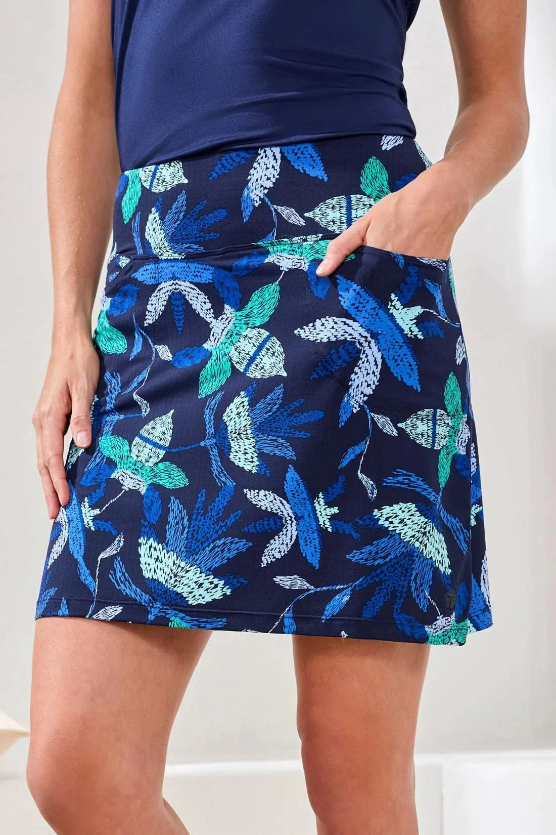 Someone stands indoors in a Tribal High Performance Skort with a navy floral print, paired with a navy top and white sneakers, one hand in their pocket.