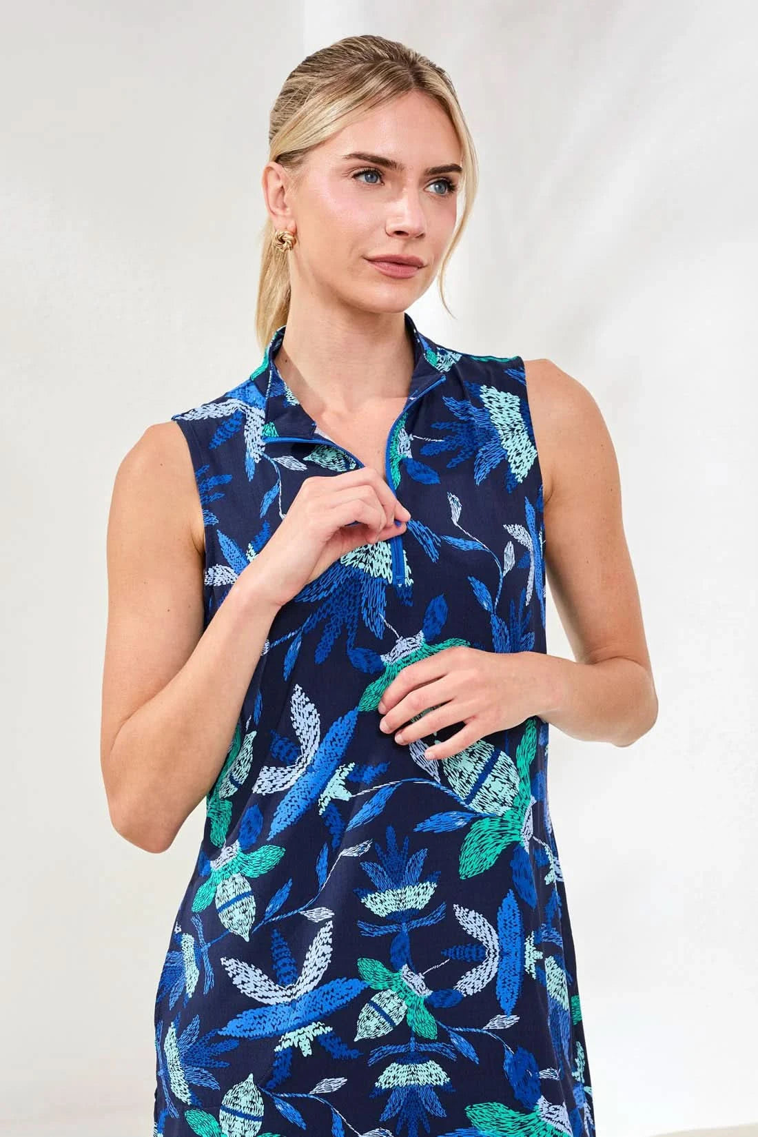 A woman with blonde hair tied back stands indoors wearing the Tribal PERFORMANCE ZIP-FRONT SPORT DRESS, a blue, sleeveless, knee-length active dress with a green and white leaf print, paired with white socks and beige sneakers.