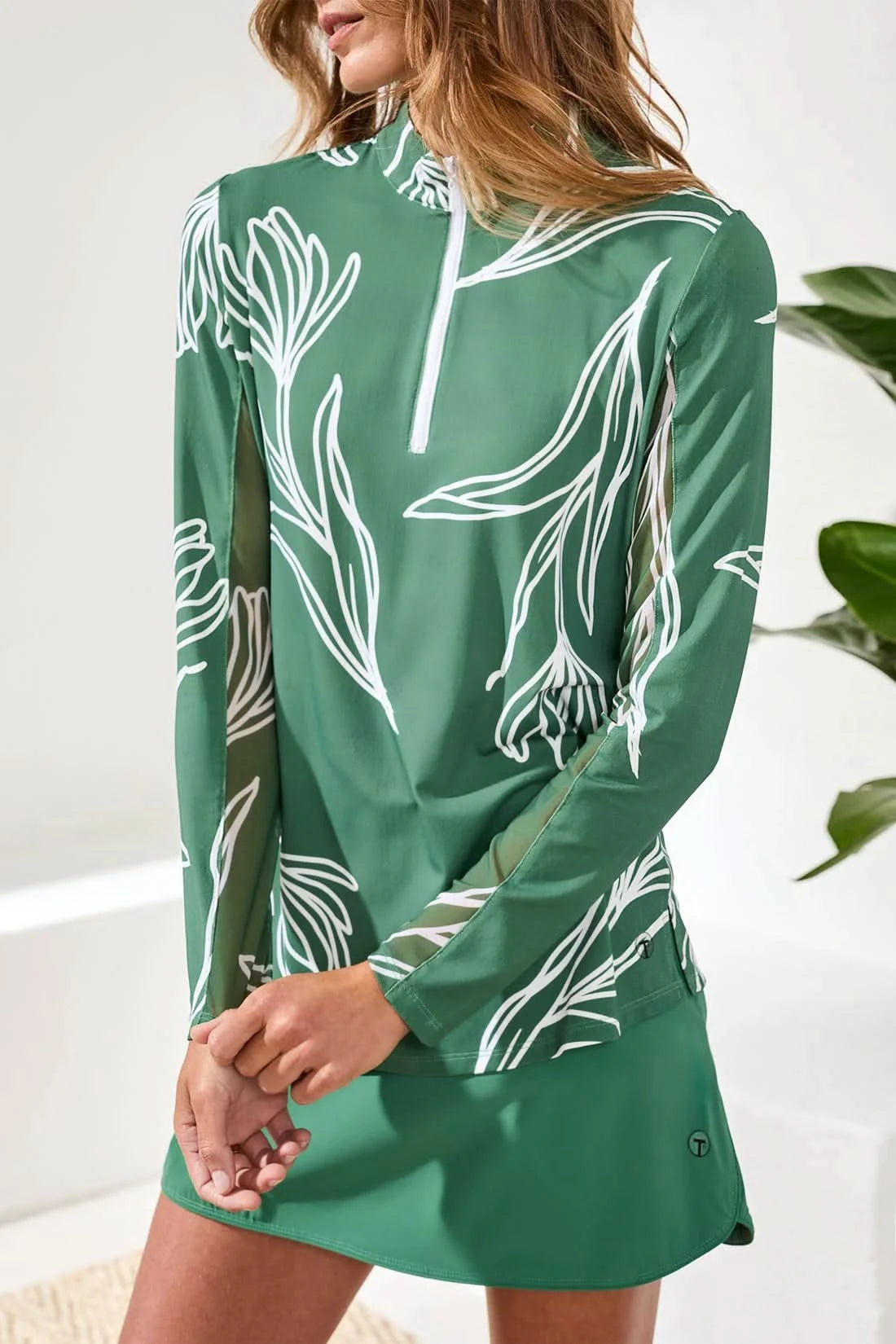 A woman stands indoors on a light woven rug, wearing Tribal's PERFORMANCE QUARTER-ZIP LONG SLEEVE in green with white leaf patterns, a matching skirt, white socks, and beige sneakers.