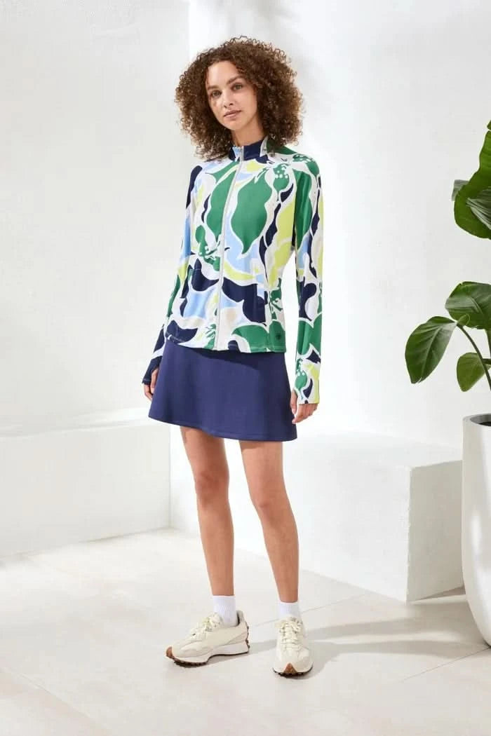 A woman wears the Tribal HIGH PERFORMANCE MESH MOCK-NECK ZIP-UP dress with a blue, green, and white leaf pattern and UPF 50+ protection, standing indoors with her hand in a pocket.