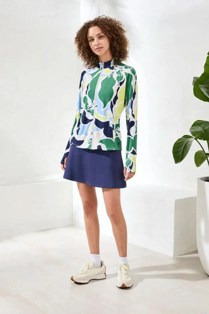 A woman wears the Tribal HIGH PERFORMANCE MESH MOCK-NECK ZIP-UP dress with a blue, green, and white leaf pattern and UPF 50+ protection, standing indoors with her hand in a pocket.