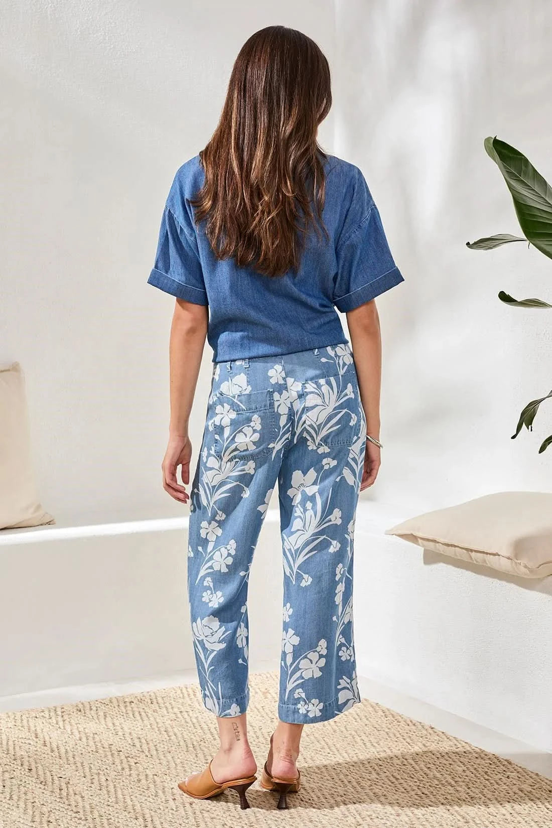 A person wearing a blue short-sleeve shirt, Tribal Flowy Wide Crop Pant, and tan heeled sandals stands indoors on a woven rug.
