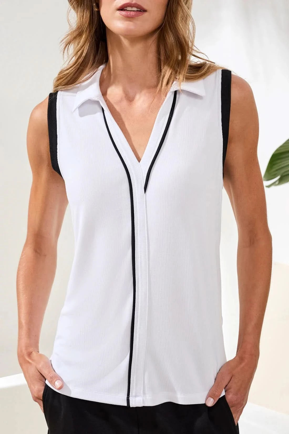 A woman stands indoors wearing the Tribal JOHNNY COLLAR MODAL TOP with black trim and loose black pants. One hand is in her pocket as she smiles slightly; a plant and cushions in the background complete this effortless summer look.