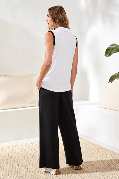 A woman stands indoors wearing the Tribal JOHNNY COLLAR MODAL TOP with black trim and loose black pants. One hand is in her pocket as she smiles slightly; a plant and cushions in the background complete this effortless summer look.