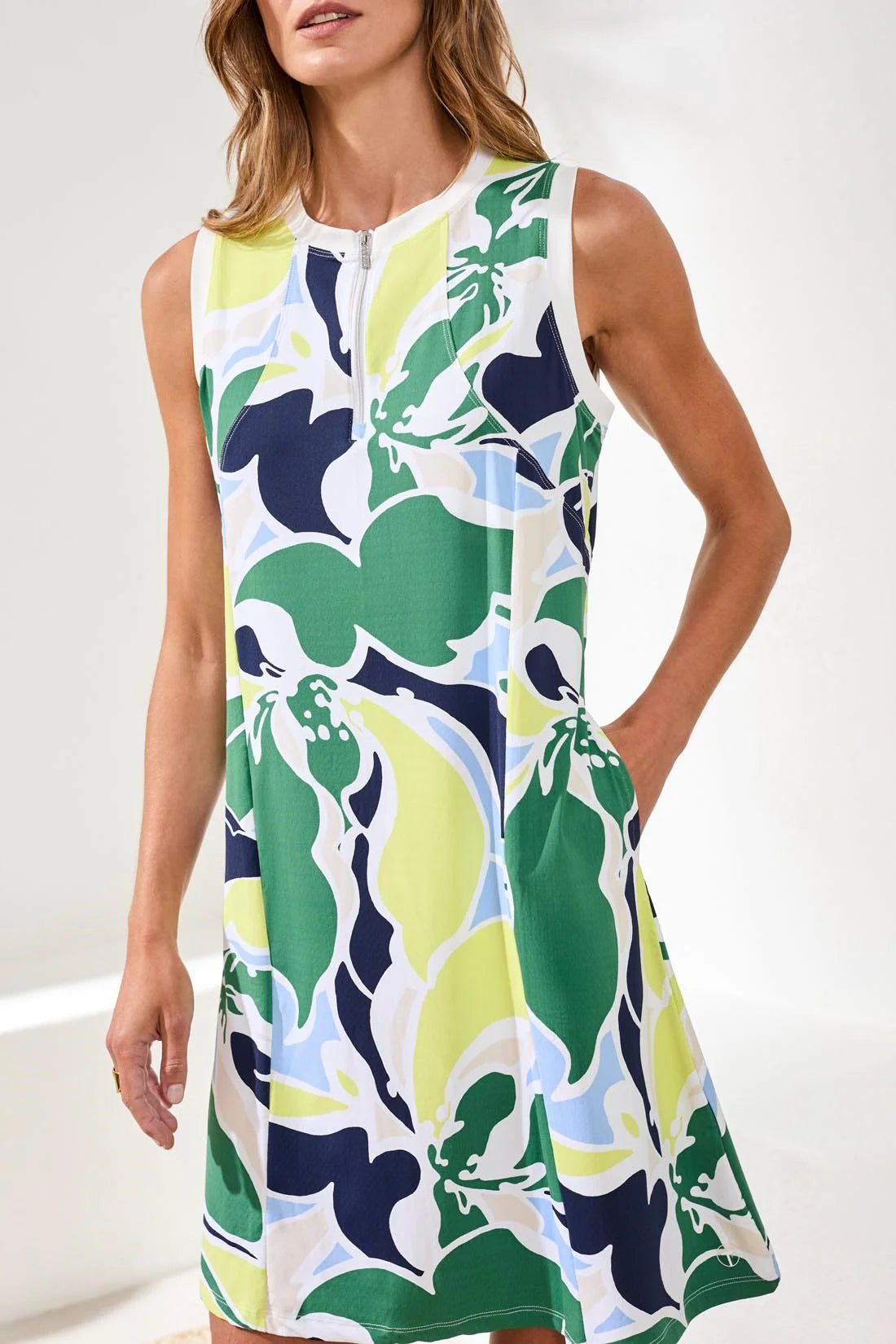 A woman smiles indoors wearing the Tribal HIGH PERFORMANCE SPORT DRESS, a sleeveless, knee-length crewneck with colorful floral print and pockets, paired with white sneakers.