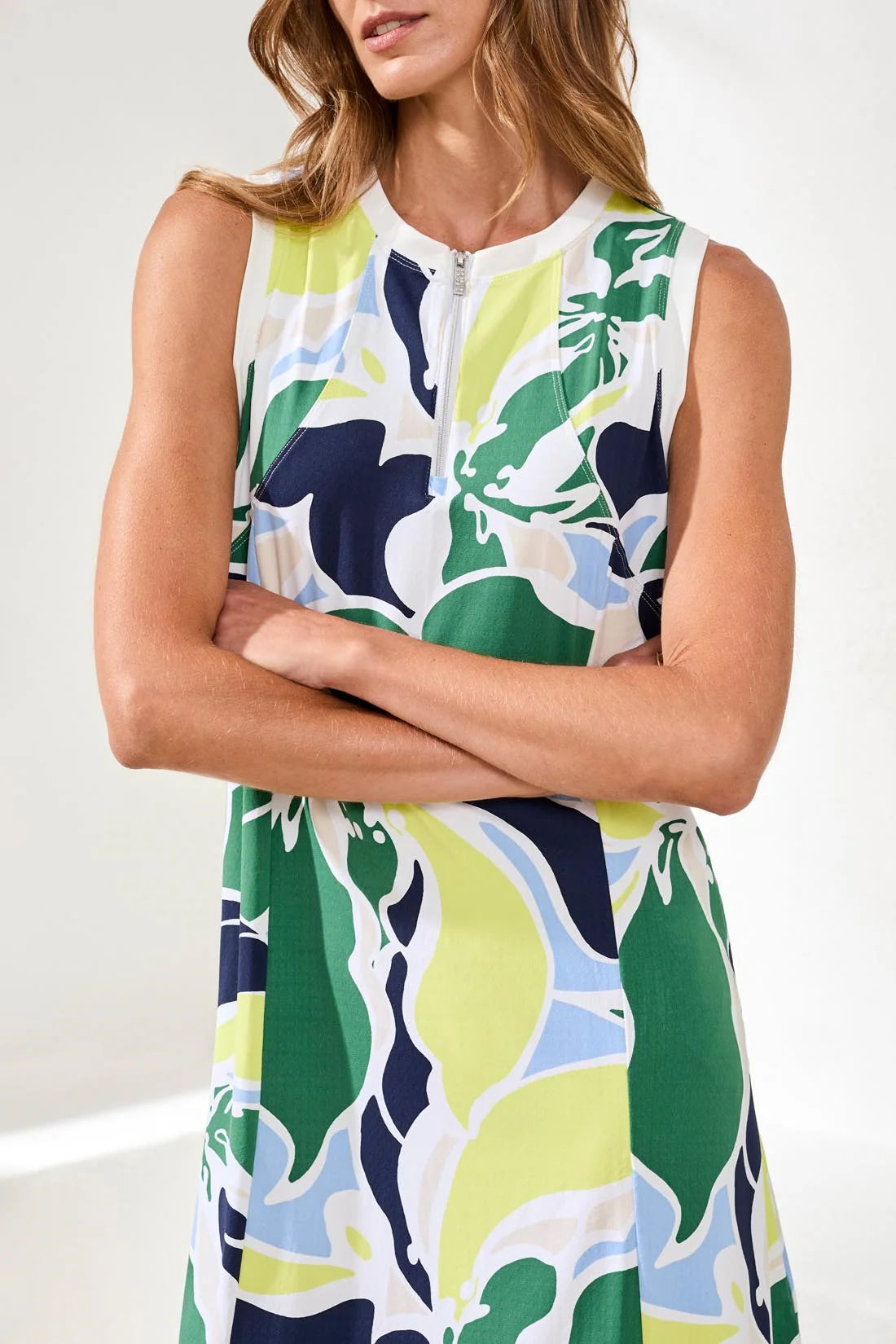 A woman smiles indoors wearing the Tribal HIGH PERFORMANCE SPORT DRESS, a sleeveless, knee-length crewneck with colorful floral print and pockets, paired with white sneakers.
