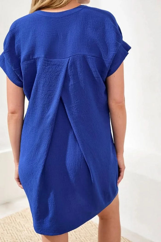 A woman stands indoors wearing the Tribal TUNIC SHIFT DRESS—a relaxed fit blue textured dress—with gold sandals and one hand in her pocket. A potted plant is seen in the background.